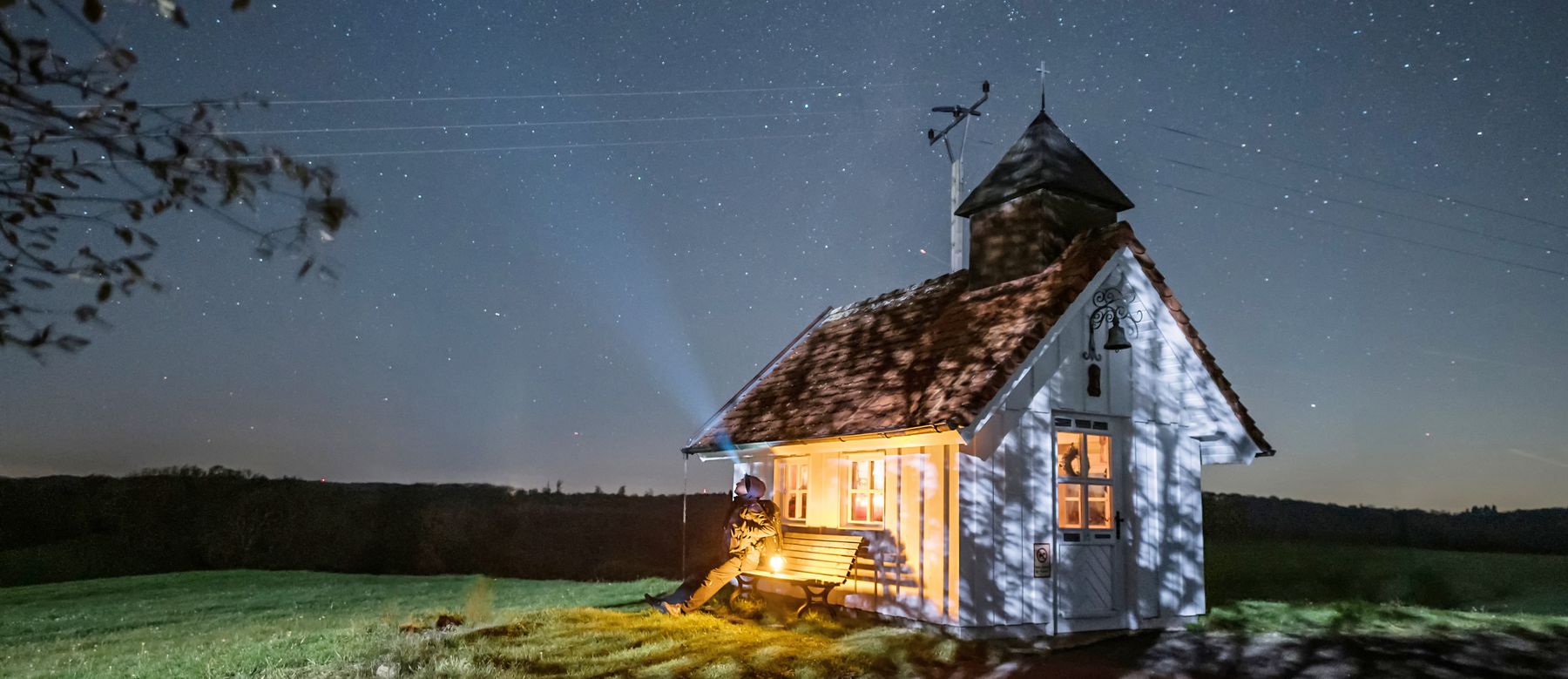 A person sits on a bench against the side of a small illuminated chapel under a starry night sky.